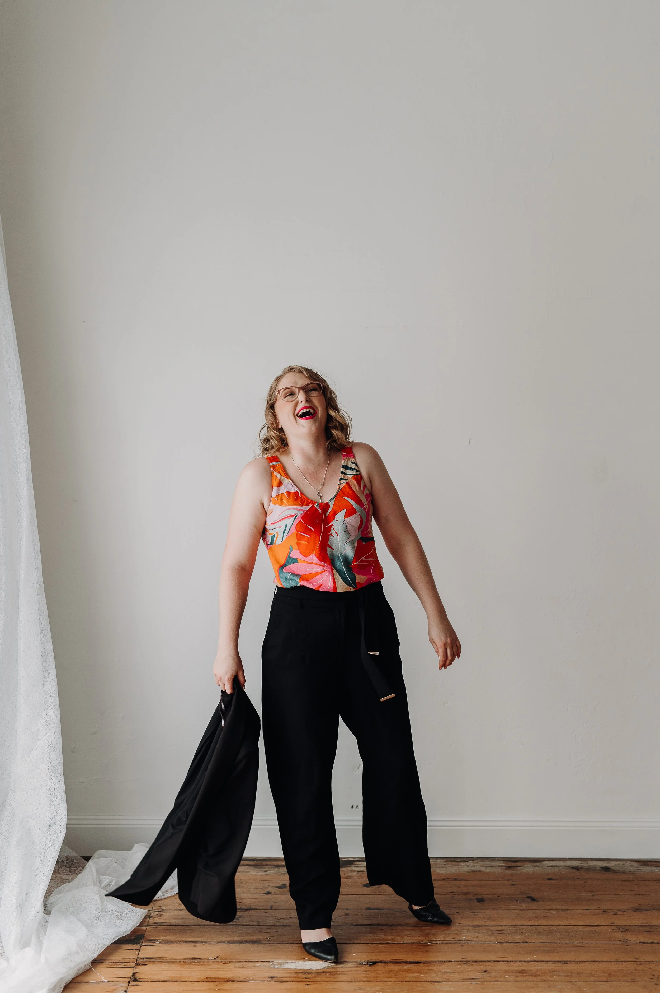 Woman with curly hair, wearing glasses, a colorful floral top, and black pants, is laughing and holding a black jacket in her left hand, standing on a wooden floor against a plain white wall.