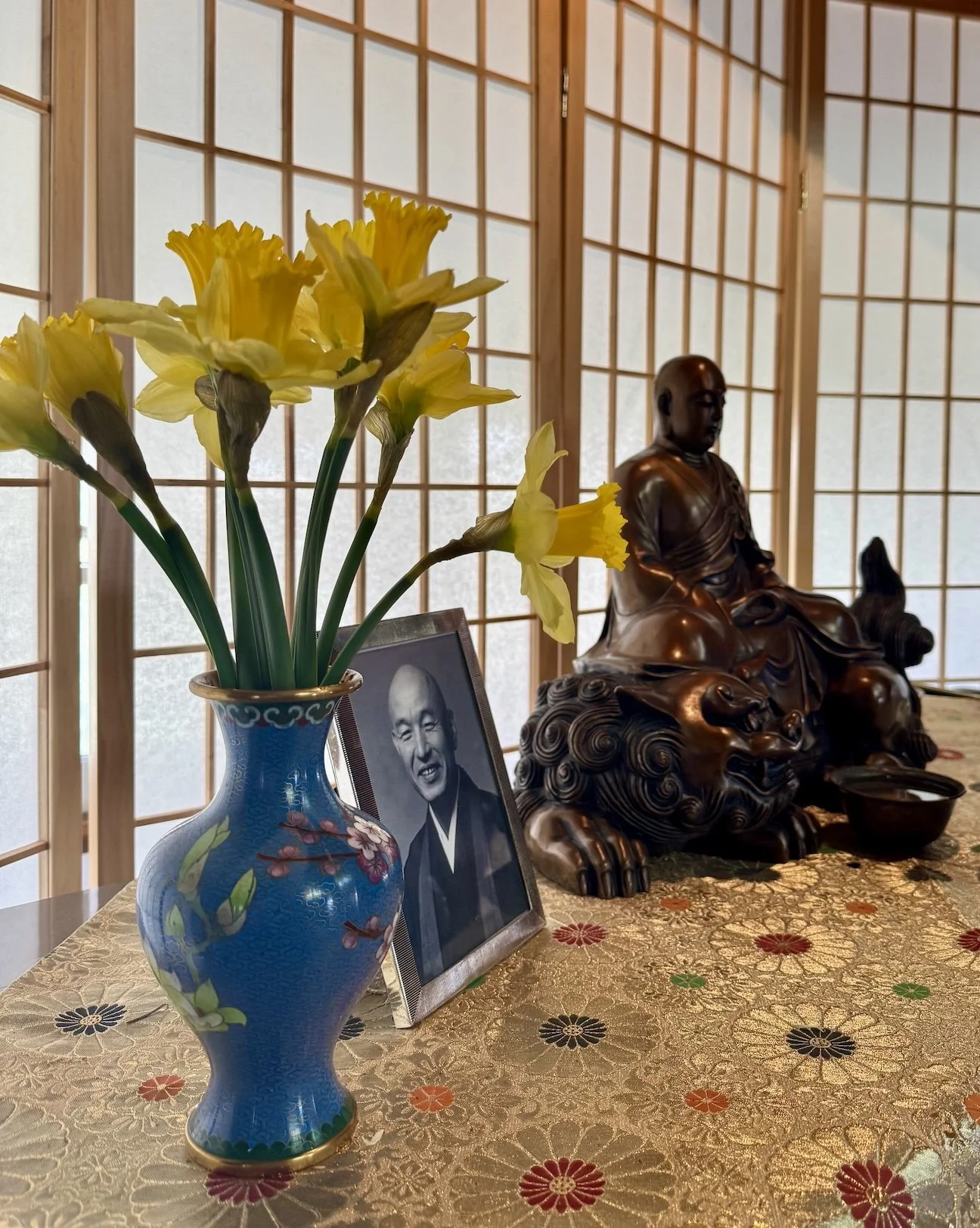 A still life image of a blue vase with yellow daffodils, a black-and-white portrait photograph of an elderly man, a Buddhist statue, and a patterned tablecloth with floral designs, set against a background of shoji screens.