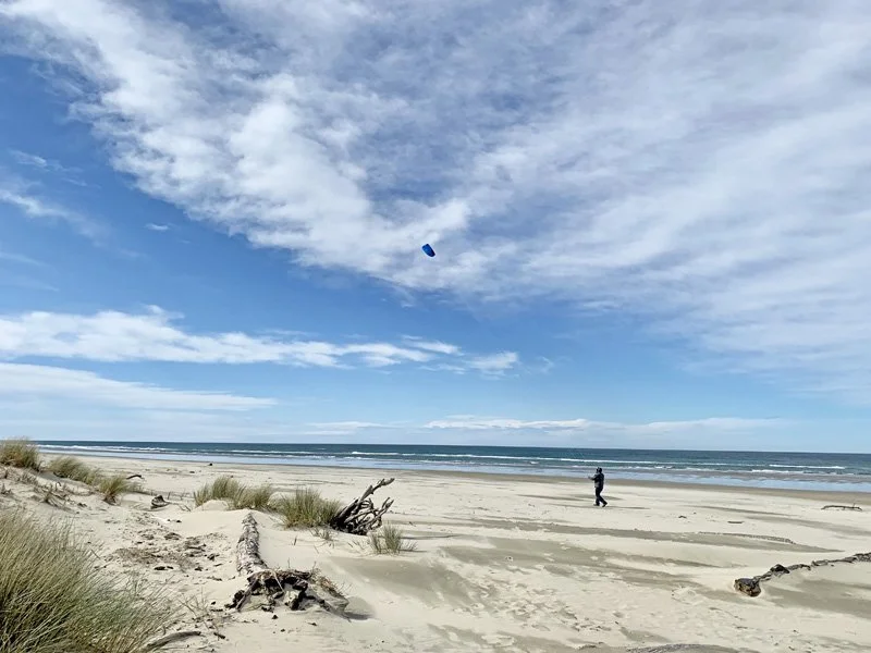 A person flying a kite on a sandy beach with grass patches, driftwood, and a blue sky with clouds