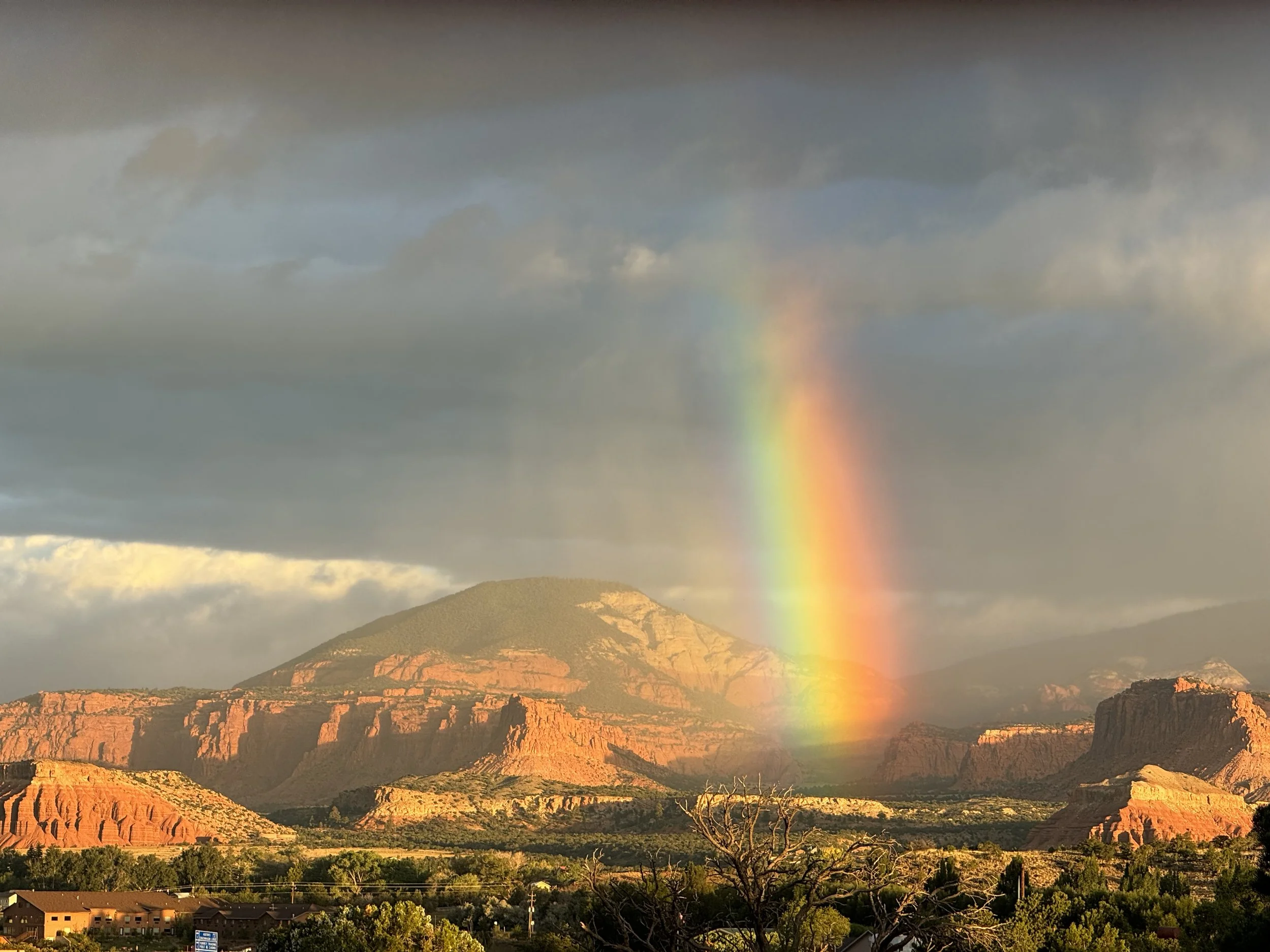 A rainbow over red rock mesas and mountains in a desert landscape, with clouds and dark sky above.
