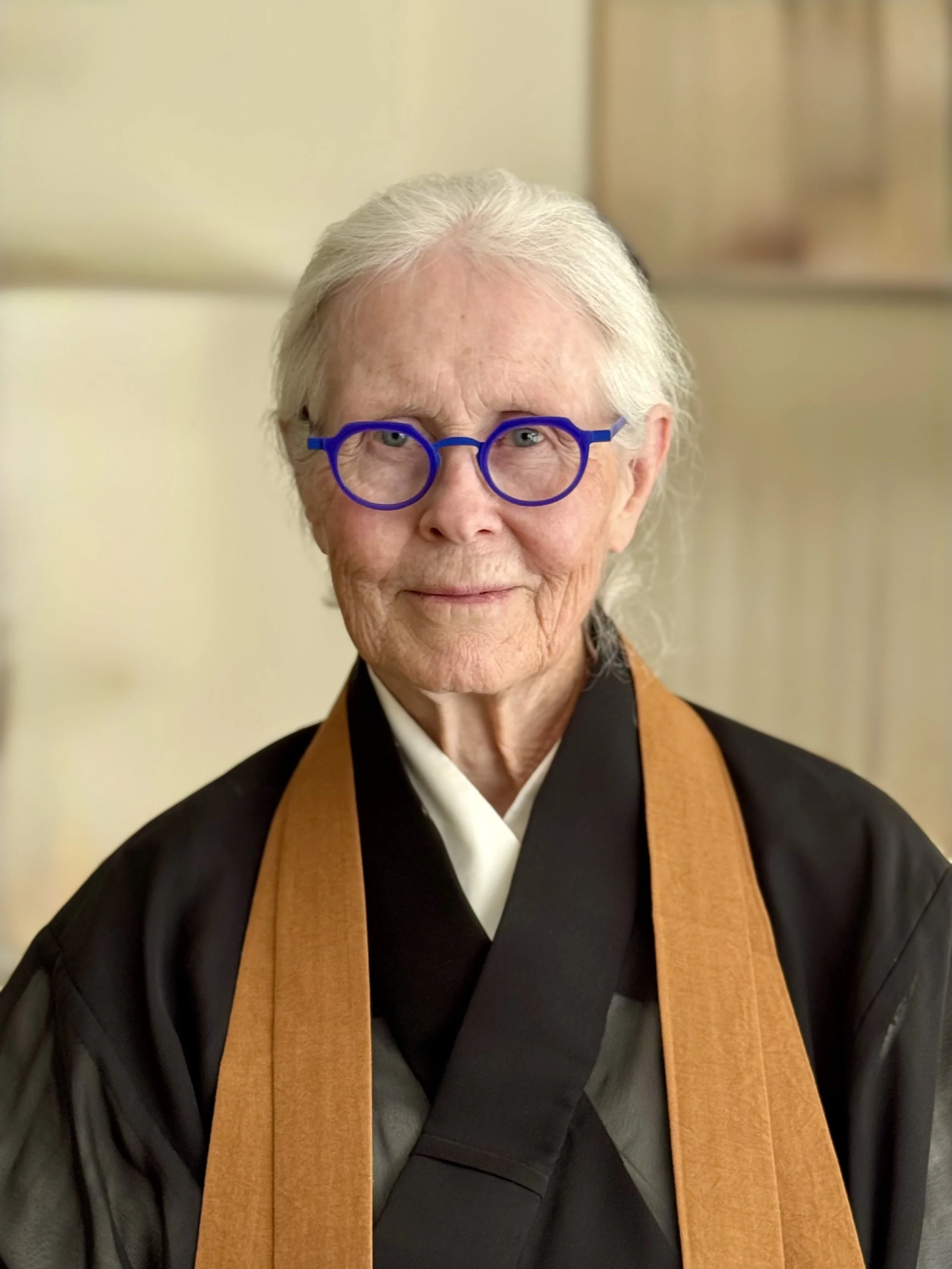 Mary Ellen Seien Roshi Zen teacher based in Salt Lake City, white hair and blue glasses, wearing traditional robes, sitting indoors with a warm expression.