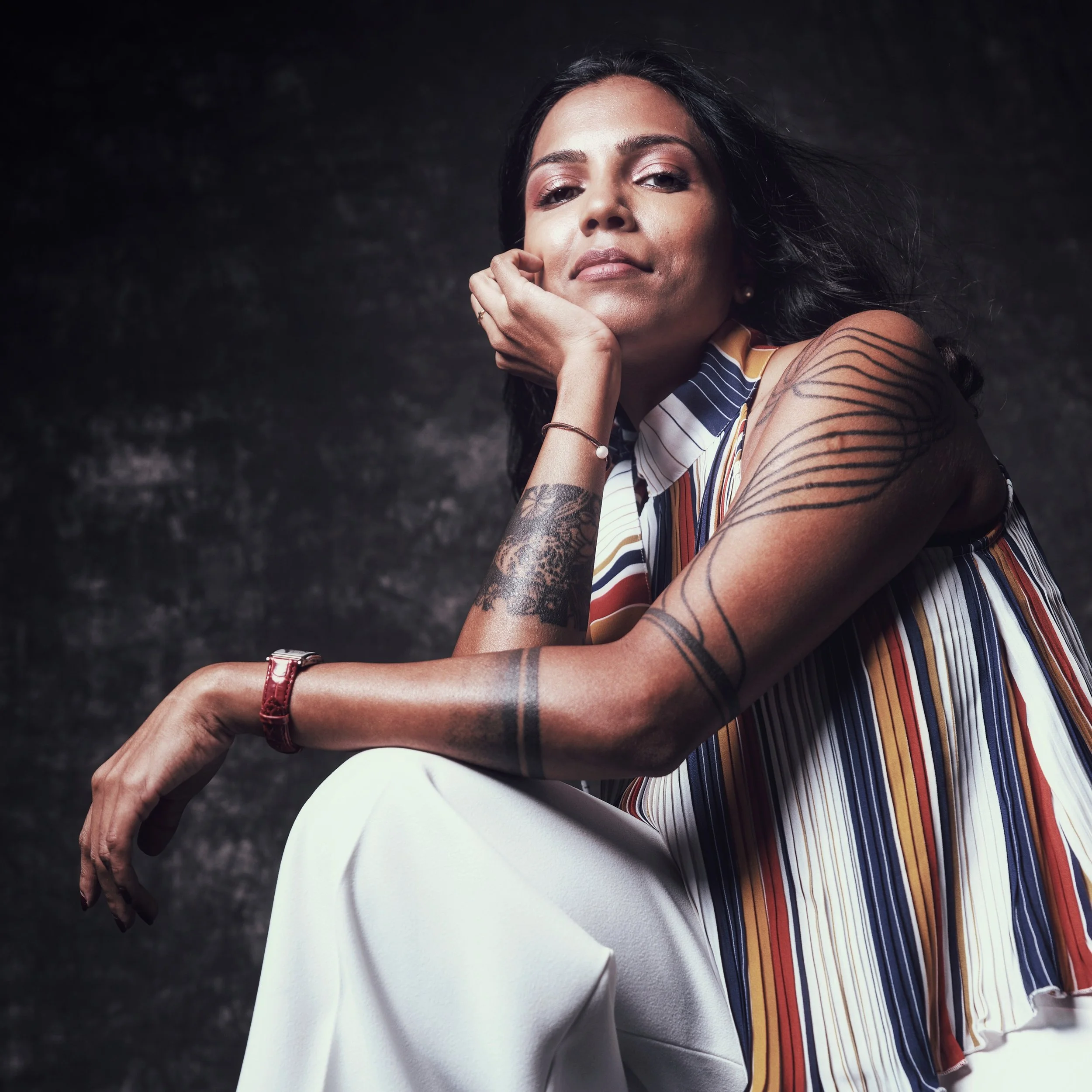 A woman with dark hair and tattoos on her arm, sitting against a dark background, wearing a striped sleeveless top and white pants.