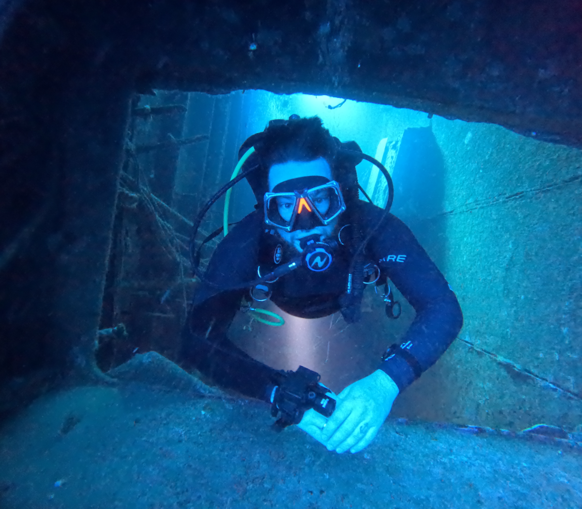 A scuba diver underwater wearing a black wetsuit, mask, and breathing apparatus, swimming through a rectangular underwater tunnel surrounded by dark rocks and blue water.