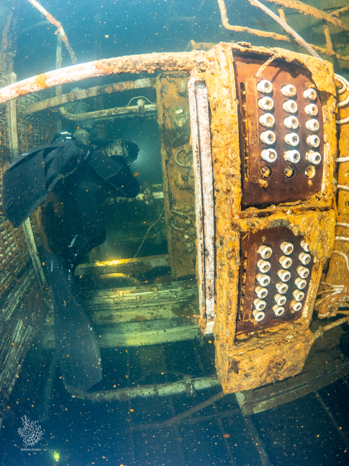 A diver explores a sunken Zenobia with rusted metal structures and wiring, illuminated by a flashlight.