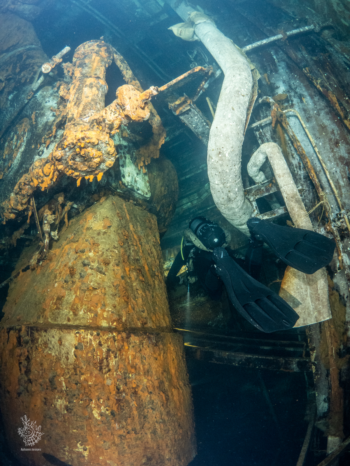 Underwater scuba diver exploring a sunken, rusted shipwreck with large pipes and machinery.