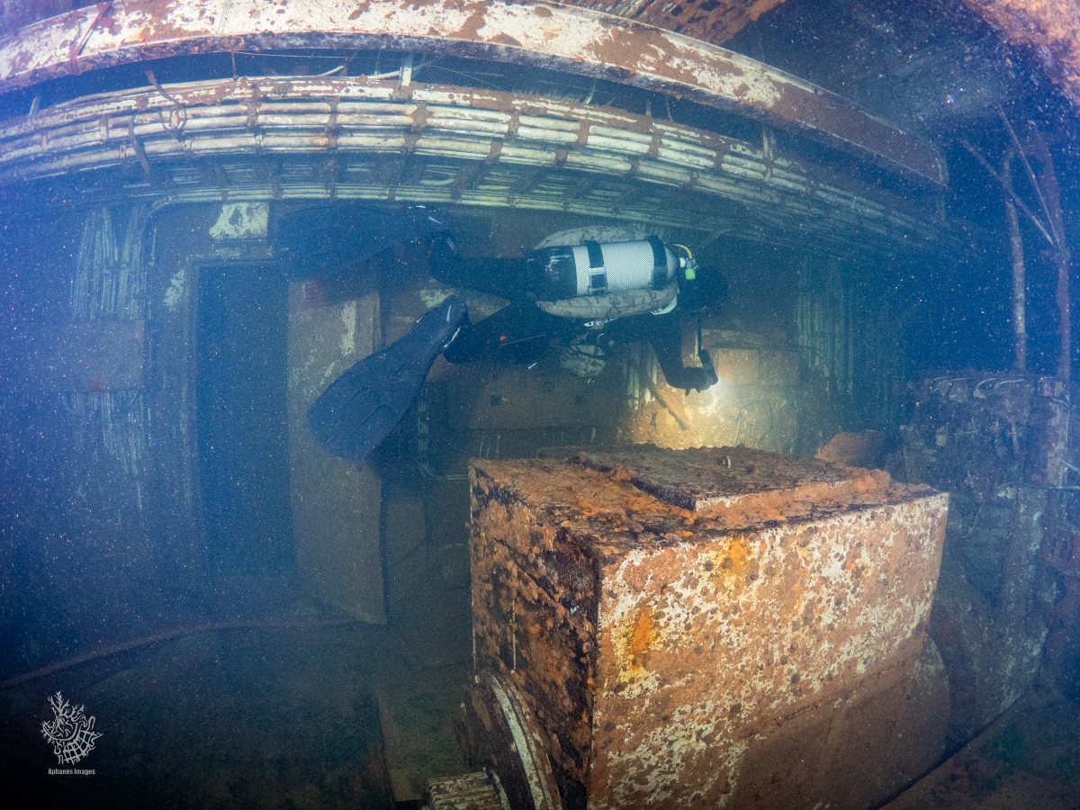 A scuba diver exploring a rusted, historic underwater shipwreck with a large rectangular cargo crate.