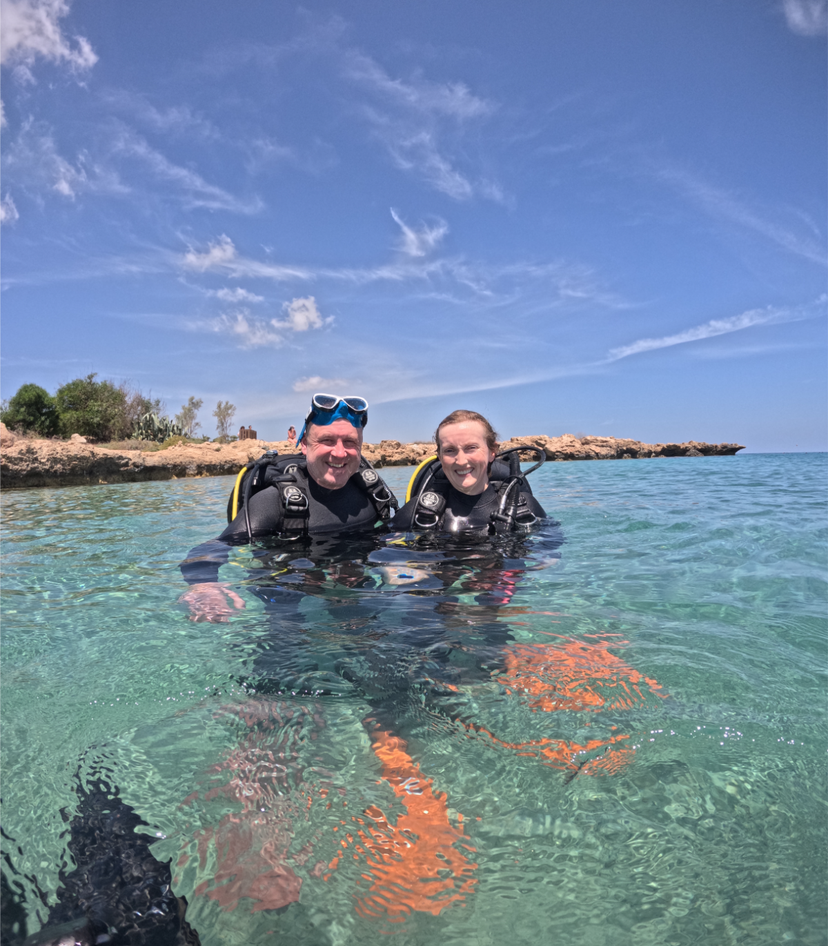 Two scuba divers in black wetsuits smiling in clear ocean water with rocky shoreline and blue sky in the background.