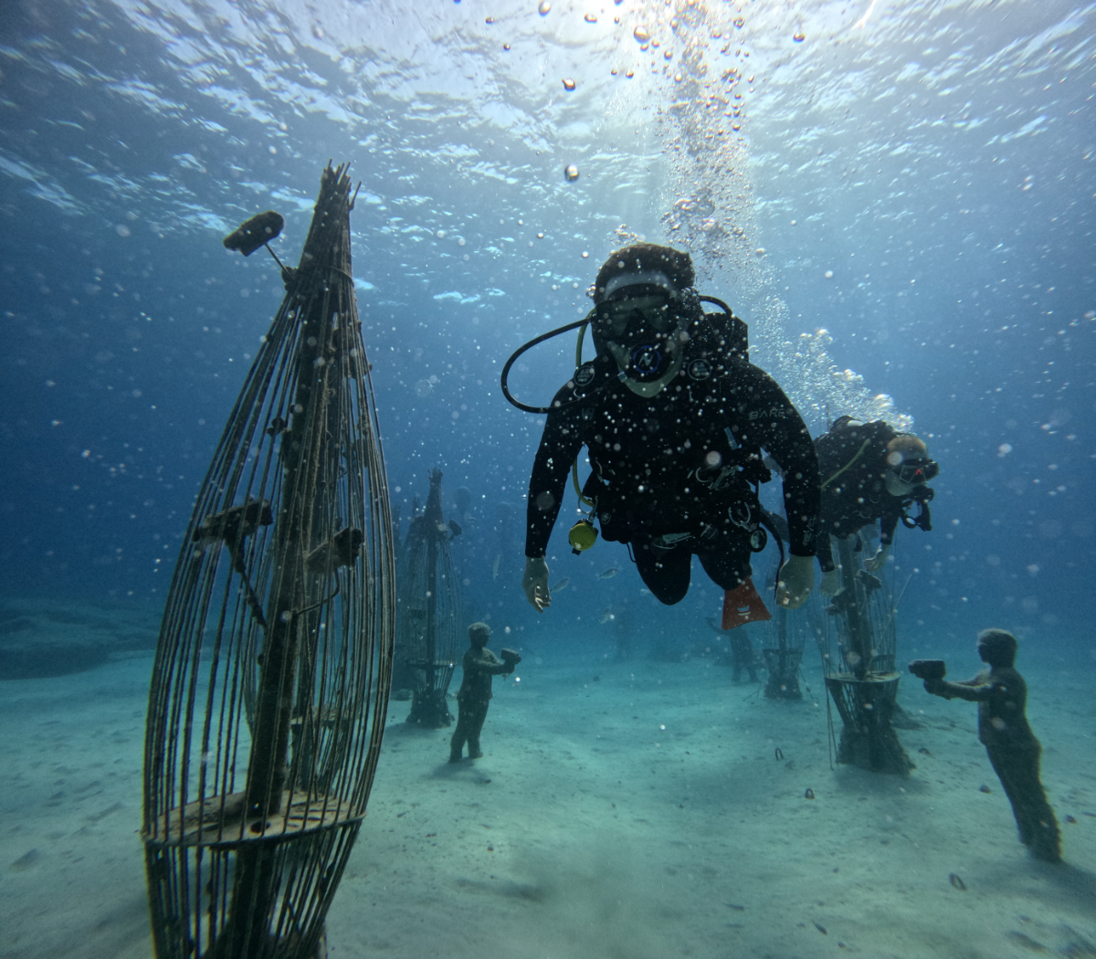 Scuba divers exploring underwater artifacts, including an submerged cage with metal bars and fish, with children observing on the sandy ocean floor and sunlight filtering through the water.