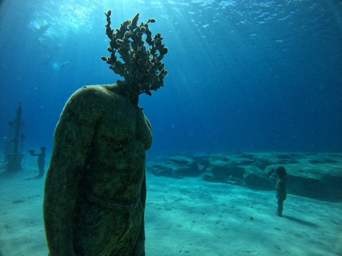 Underwater sculpture of a person with a head made of coral or brain coral, standing on a sandy ocean floor with other divers and a small child in the background, illuminated by sunlight.