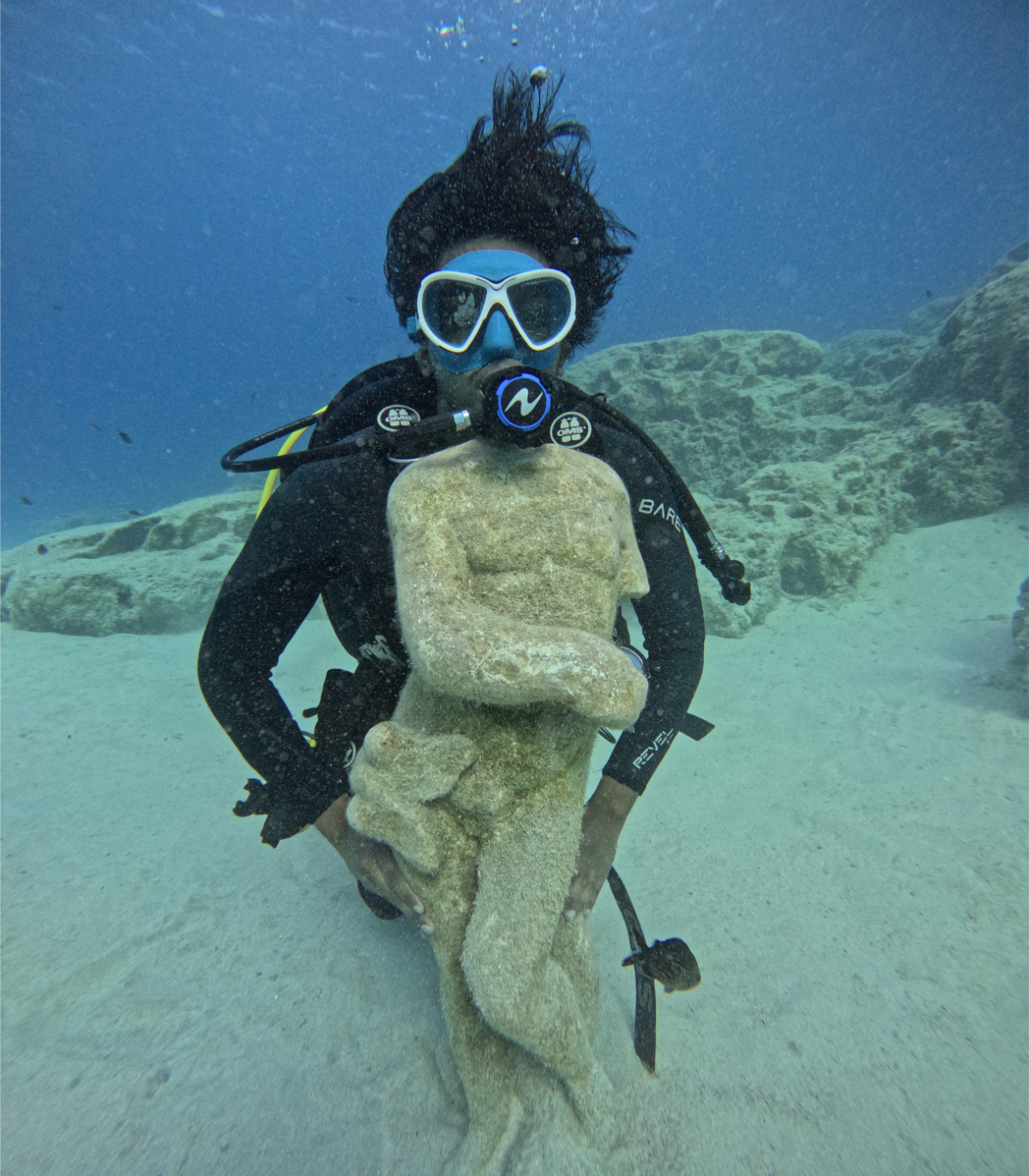 An underwater scene with a scuba diver wearing a mask and breathing apparatus, holding a large coral formation resembling a face with a protruding tongue on the sandy ocean floor with rocks and coral in the background.