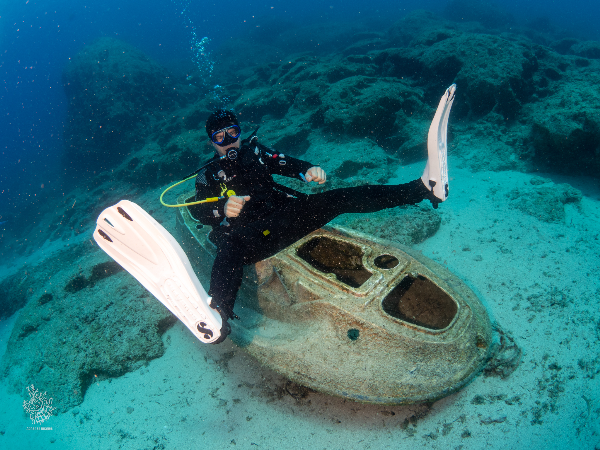 A scuba diver sitting on a sunken, rusty boat underwater, wearing a wetsuit, mask, and fins.