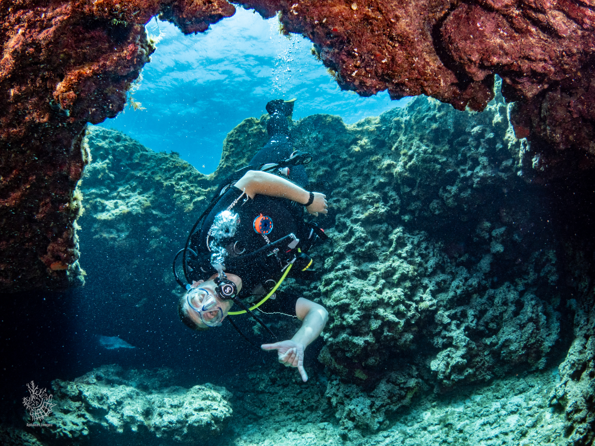 A scuba diver exploring underwater rocks and coral formations, wearing a wetsuit, mask, and breathing apparatus, waving with one hand.