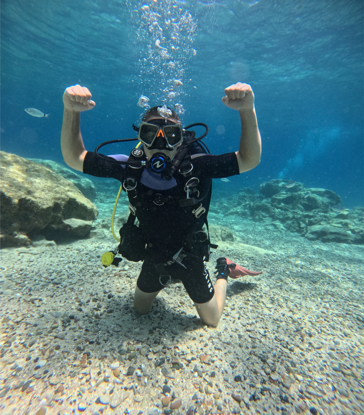 A person in scuba gear kneeling underwater on a sandy seabed with rocks, raising their arms in a flexing pose.