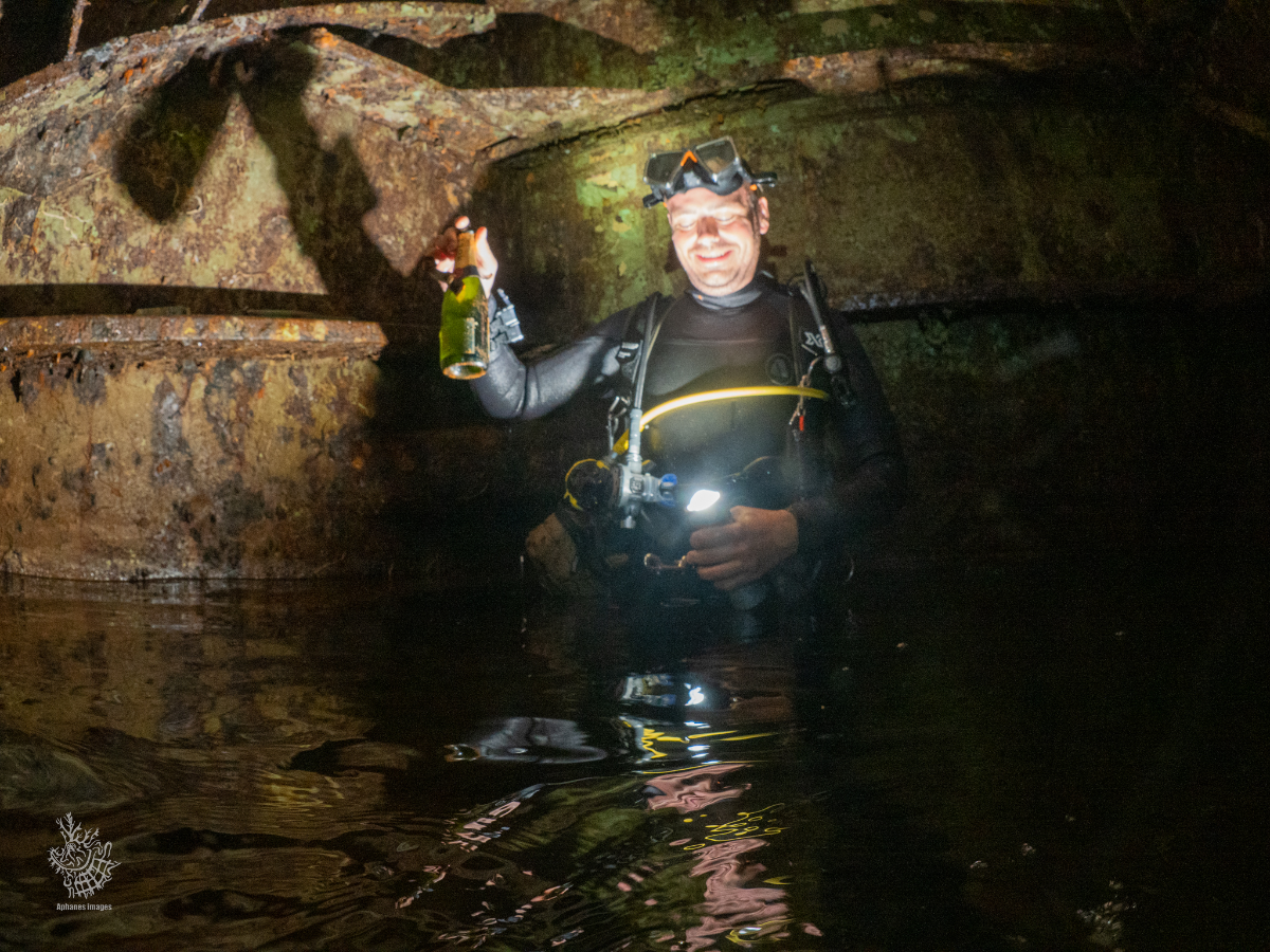 A man in scuba diving gear, wearing a headlamp and wetsuit, standing waist-deep in water inside the engine room of the Zenobia wreck smiling and holding a bottle.