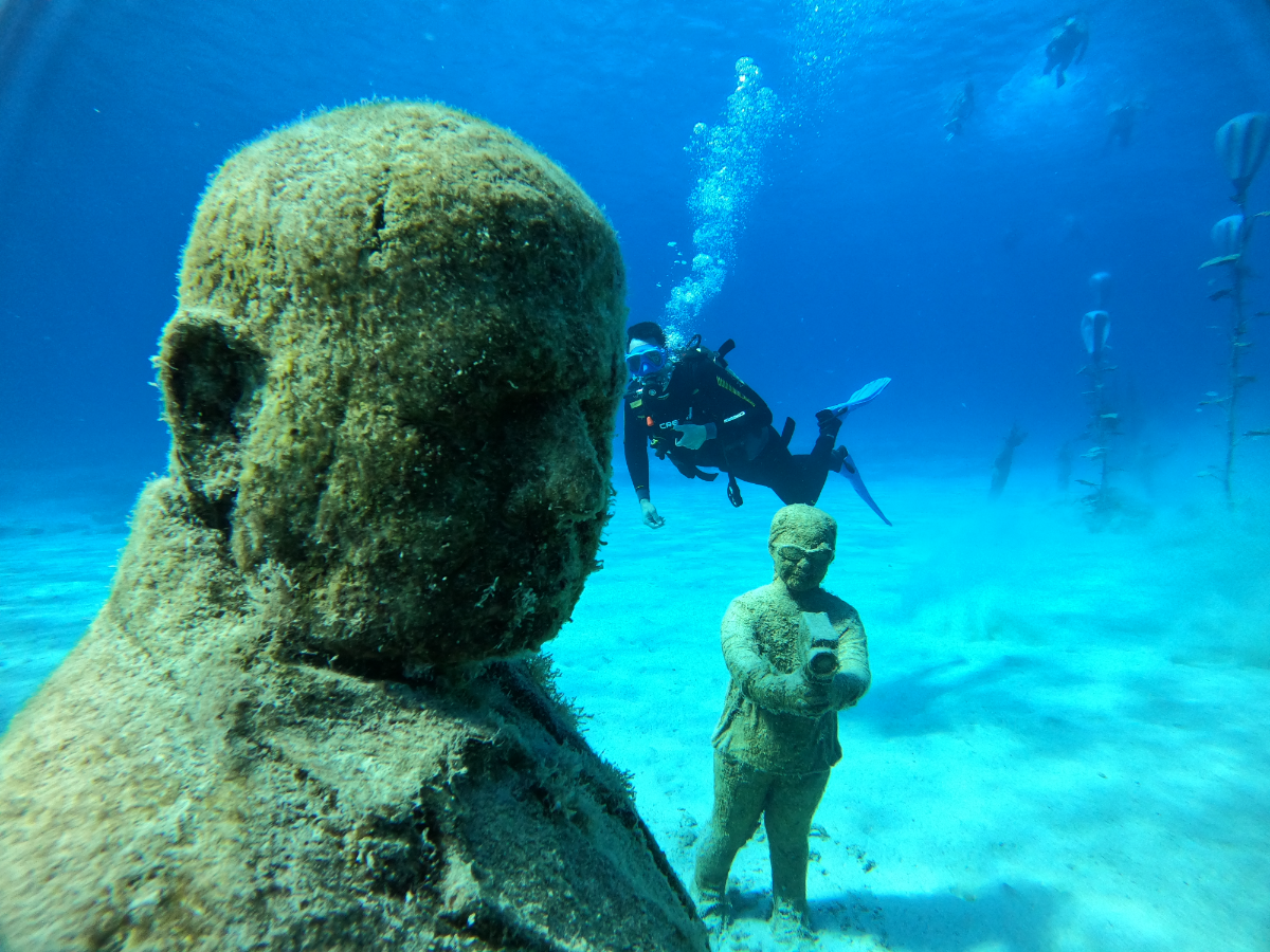 Underwater scene with a diver swimming near large stone sculptures of a child's face and a figure holding a gun, on the ocean floor with tall aquatic plants in the background.