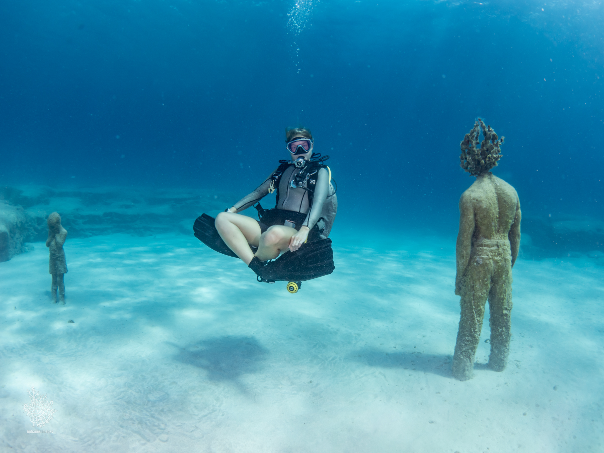 A person with scuba gear sitting on a floating platform underwater, surrounded by statues on the ocean floor.