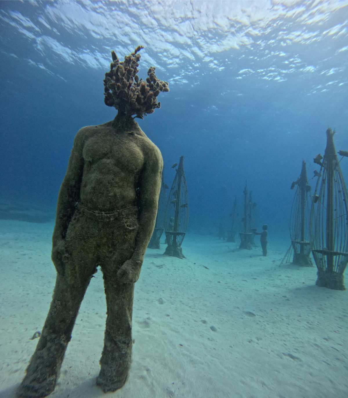Underwater sculpture of a human figure with a coral head, set on a sandy ocean floor with ghostly ships in the background.