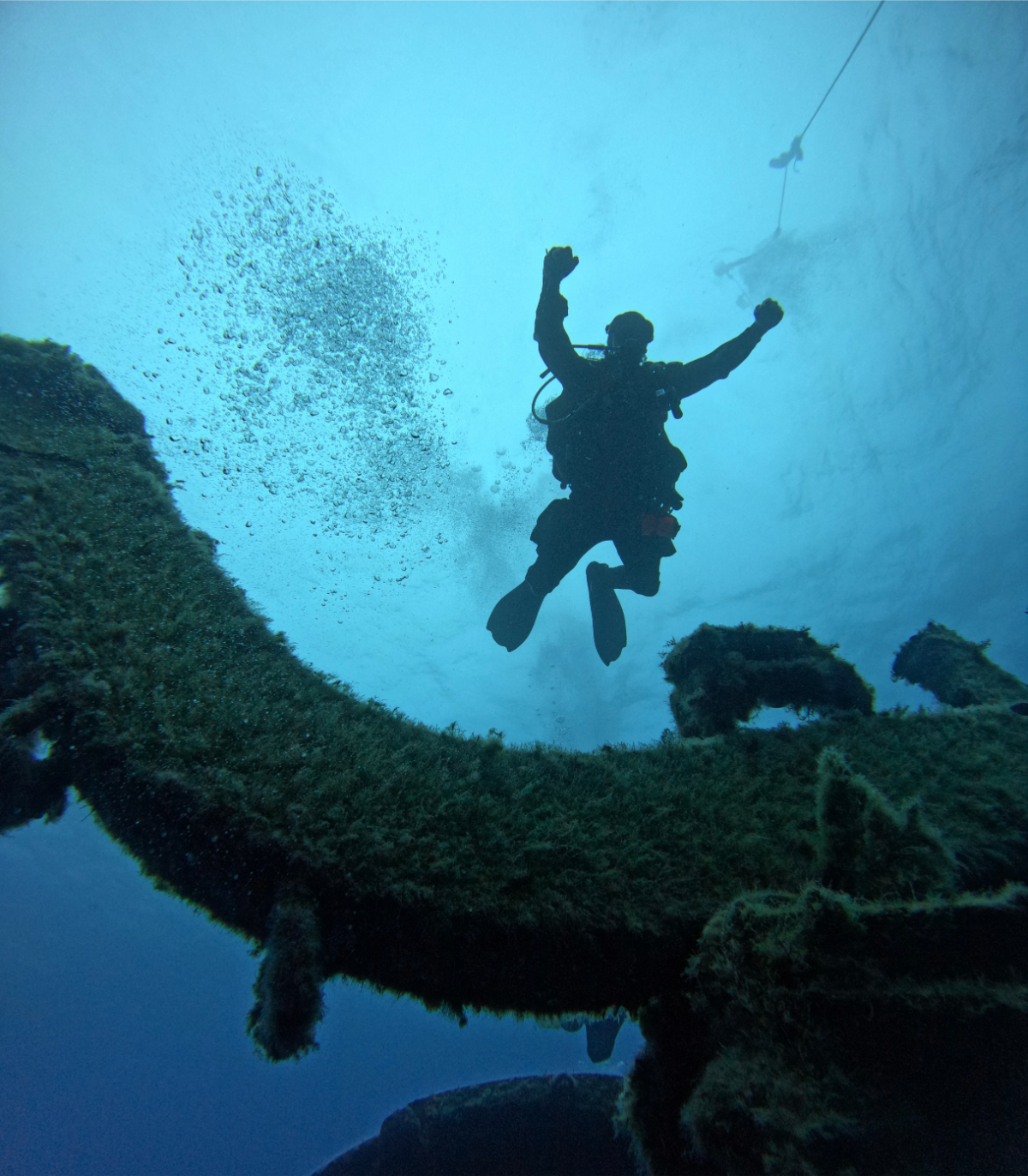 Scuba diver underwater near coral reef, bubbles, with two other divers visible in the distance.