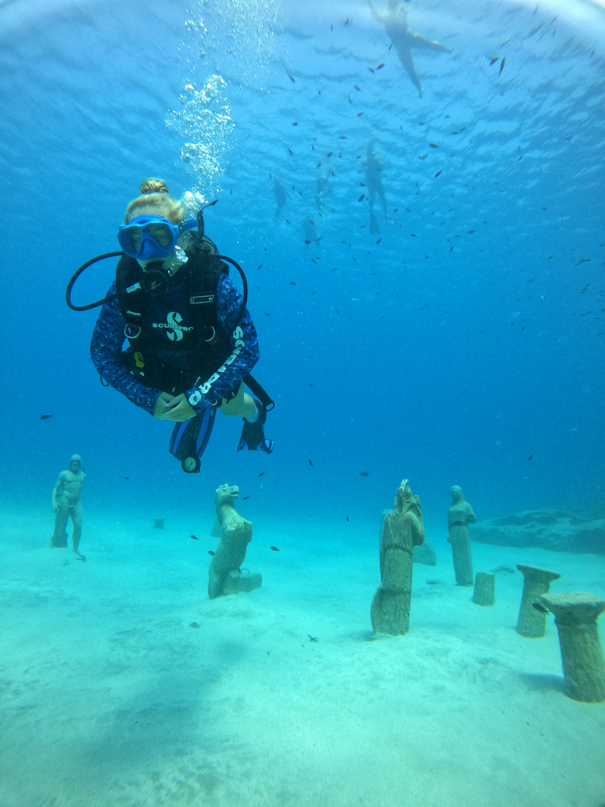A woman scuba diving underwater surrounded by fish with three totem-like wooden statues on the sandy ocean floor.