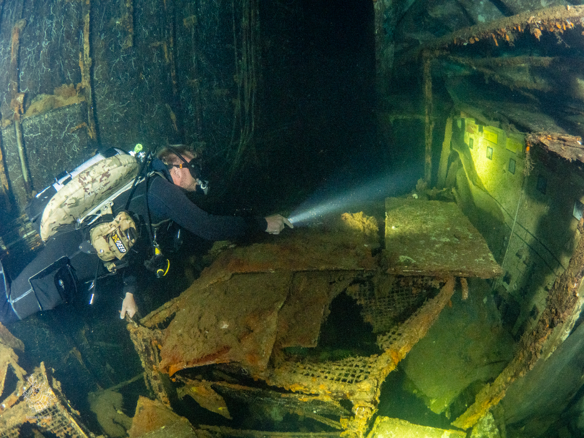 A scuba diver exploring the interior of a sunken, rusted Zenobia wreck underwater, with the diver holding a flashlight illuminating the wreckage.
