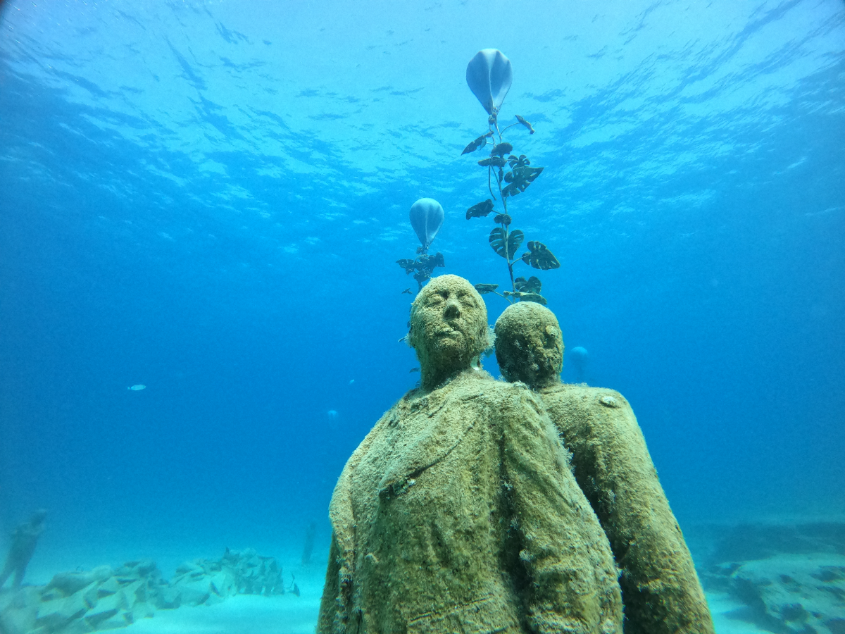 Underwater sculpture of two human figures with plant-like structures growing from their heads, set on the ocean floor.