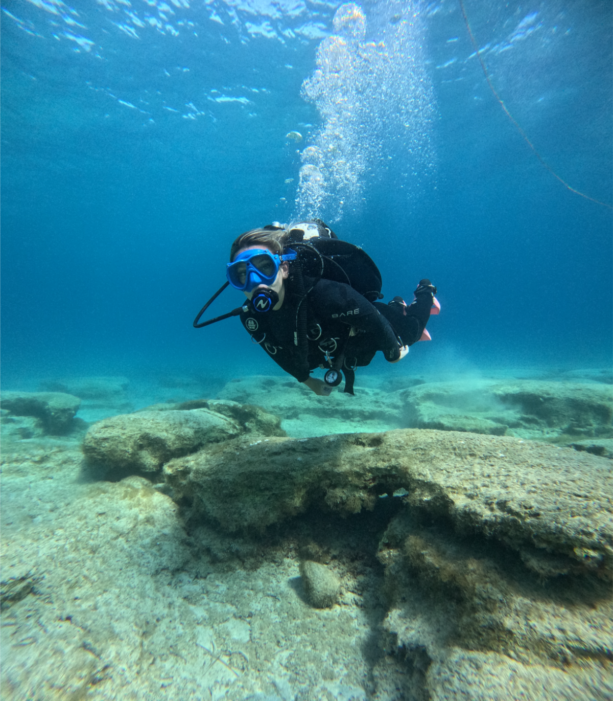 Person scuba diving underwater near rocks, wearing a wetsuit, mask, and breathing apparatus.