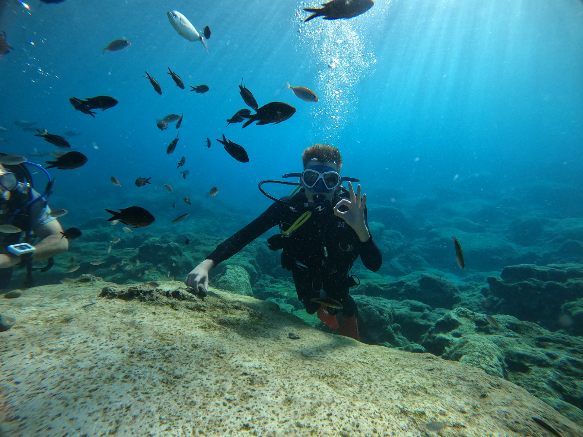 A scuba diver underwater making an OK hand gesture surrounded by fish and coral.