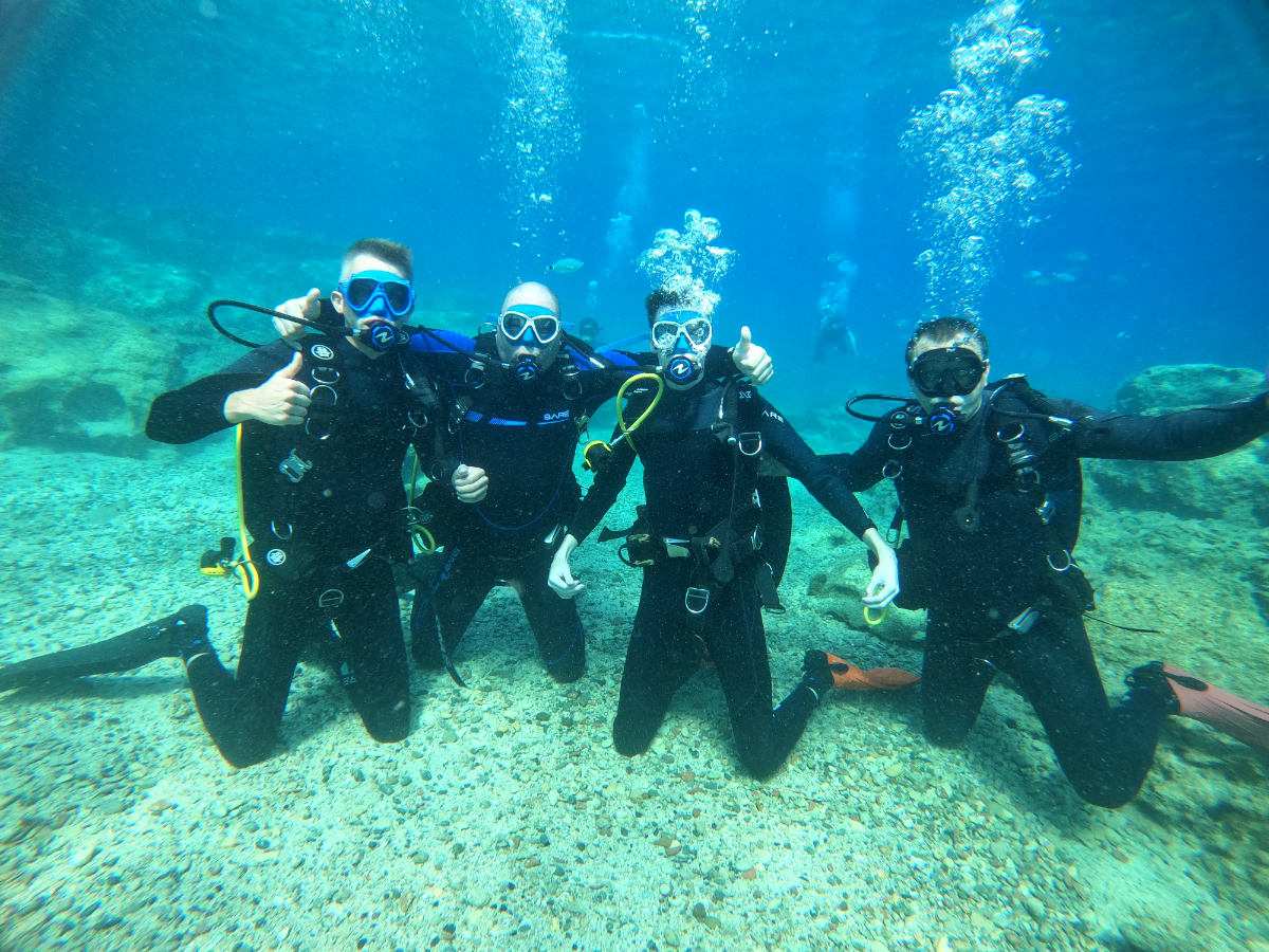 Four scuba divers underwater posing for a photo, with one giving a thumbs-up and another with arms outstretched.