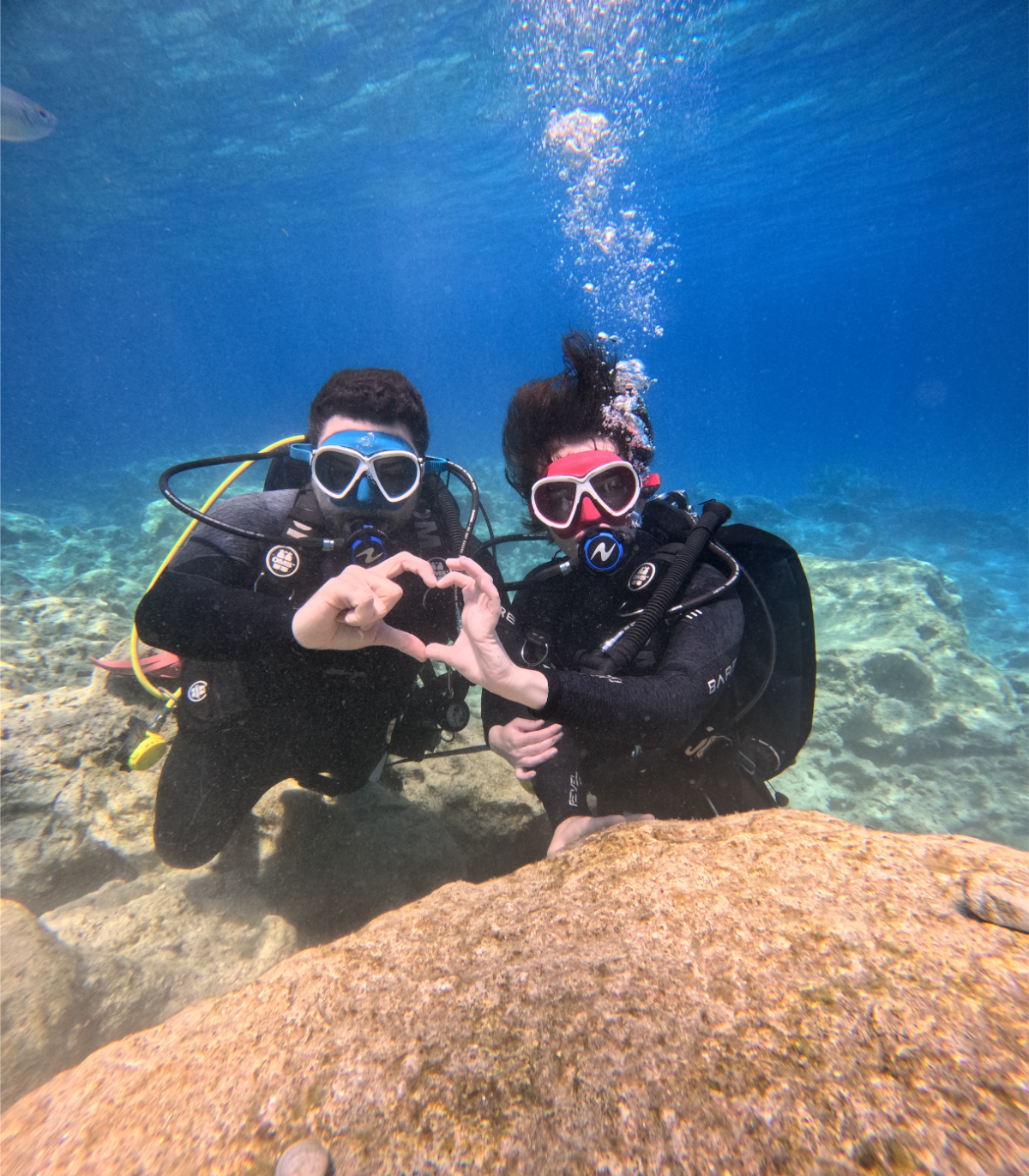 Two discover scuba divers underwater making a heart shape with their hands, wearing wetsuits, masks, and regulators, on a rocky ocean floor.