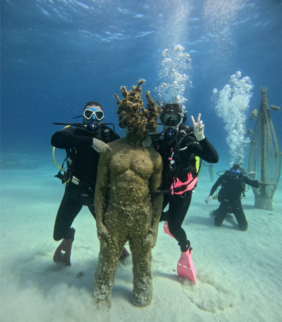 Two scuba divers posing with a sand statue of a woman on the beach underwater, giving peace signs.