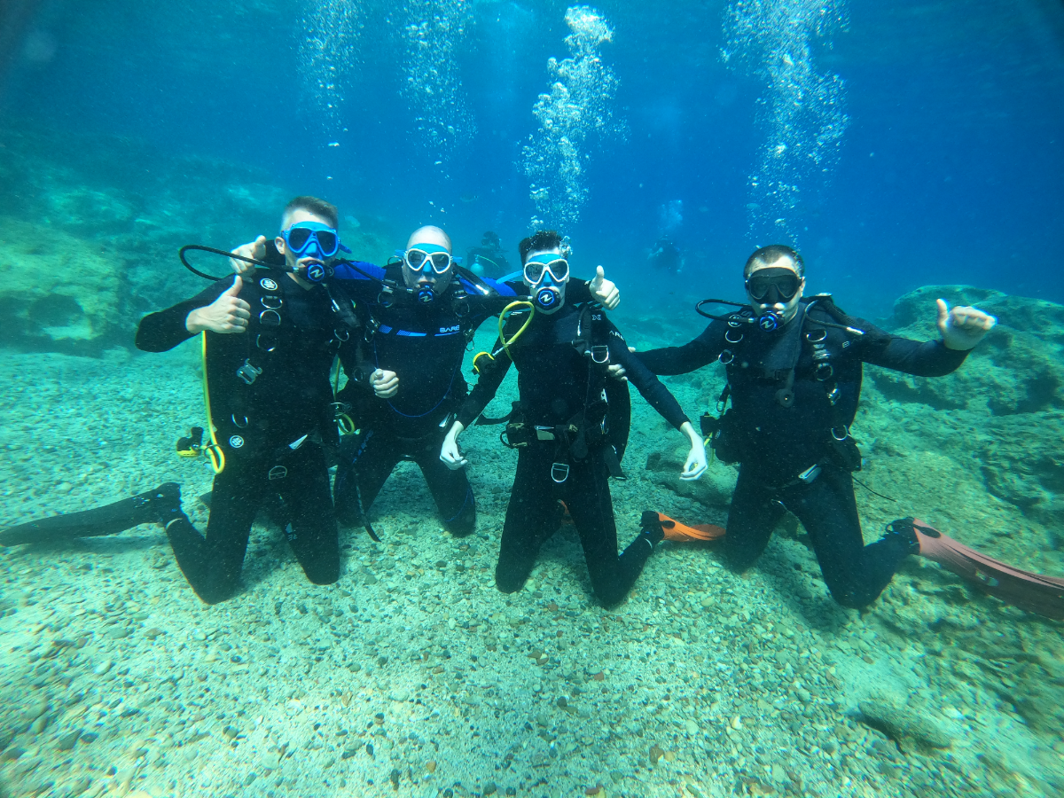 Four scuba divers underwater, kneeling on the ocean floor with rocks and sand, wearing wetsuits, masks, and breathing gear, giving thumbs up and posing for the camera.