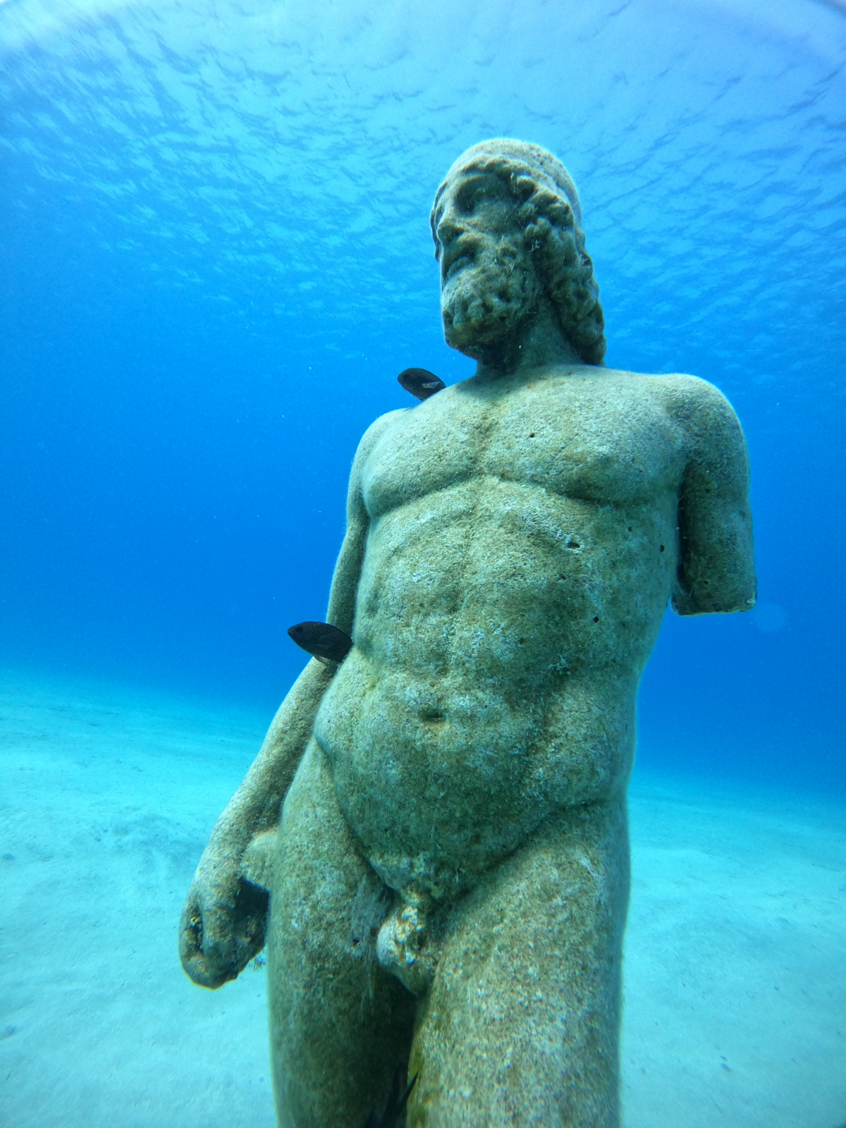Ancient Greek-style stone statue of a male figure underwater with three fish attached to it.