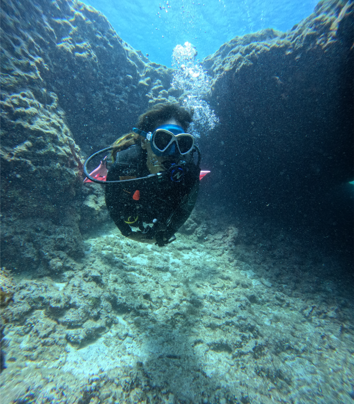 A person scuba diving underwater among rocks and coral, wearing a wetsuit, mask, and breathing apparatus, with bubbles rising to the surface.