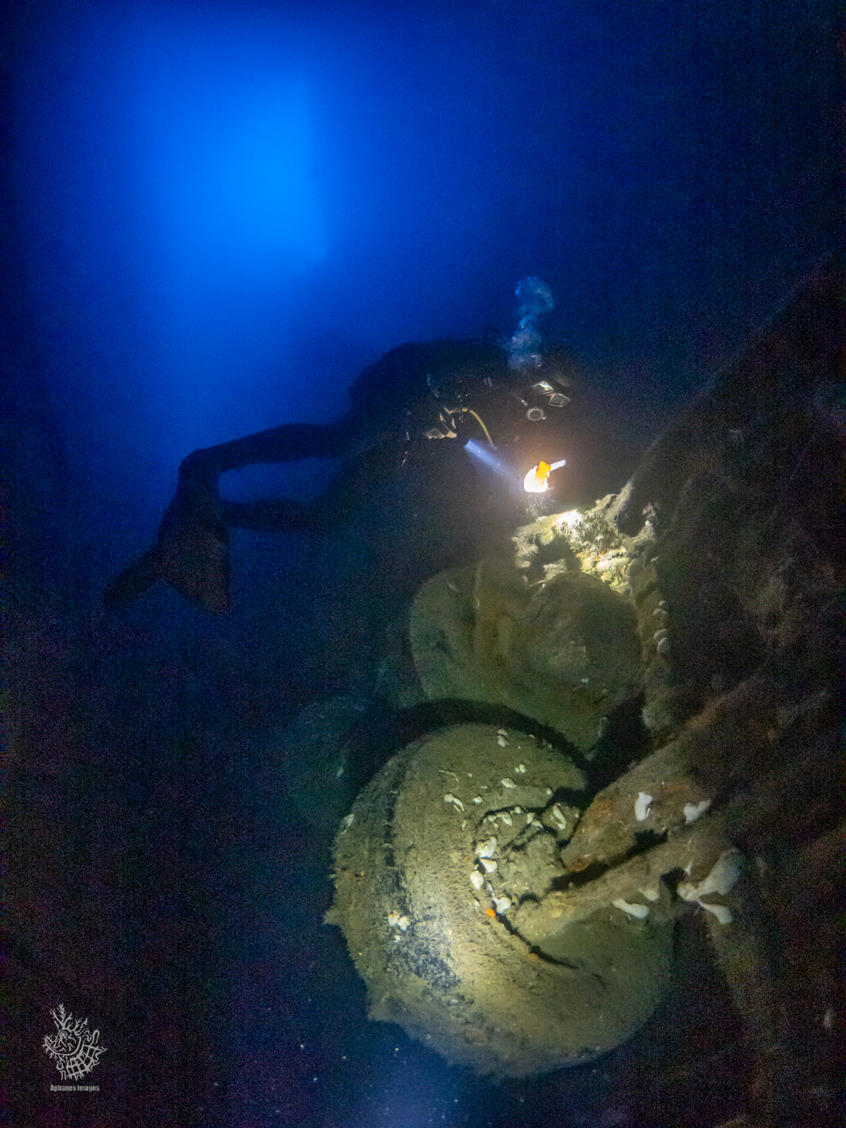A scuba diver explores a shipwreck underwater in darkness, illuminated by a flashlight.