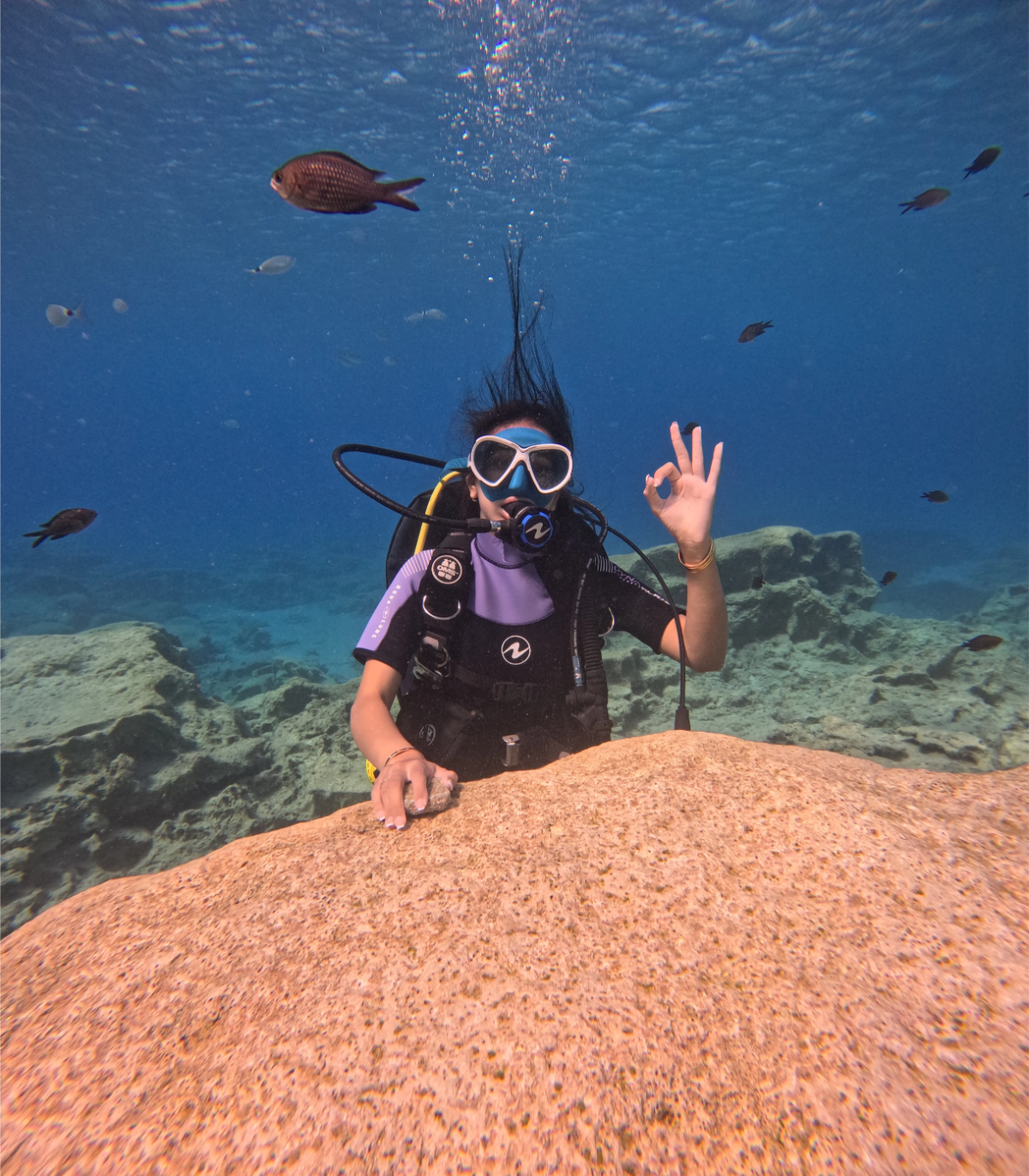 A woman scuba diving underwater, wearing a mask and wetsuit, holding onto a large rock, and making an okay sign with her fingers.
