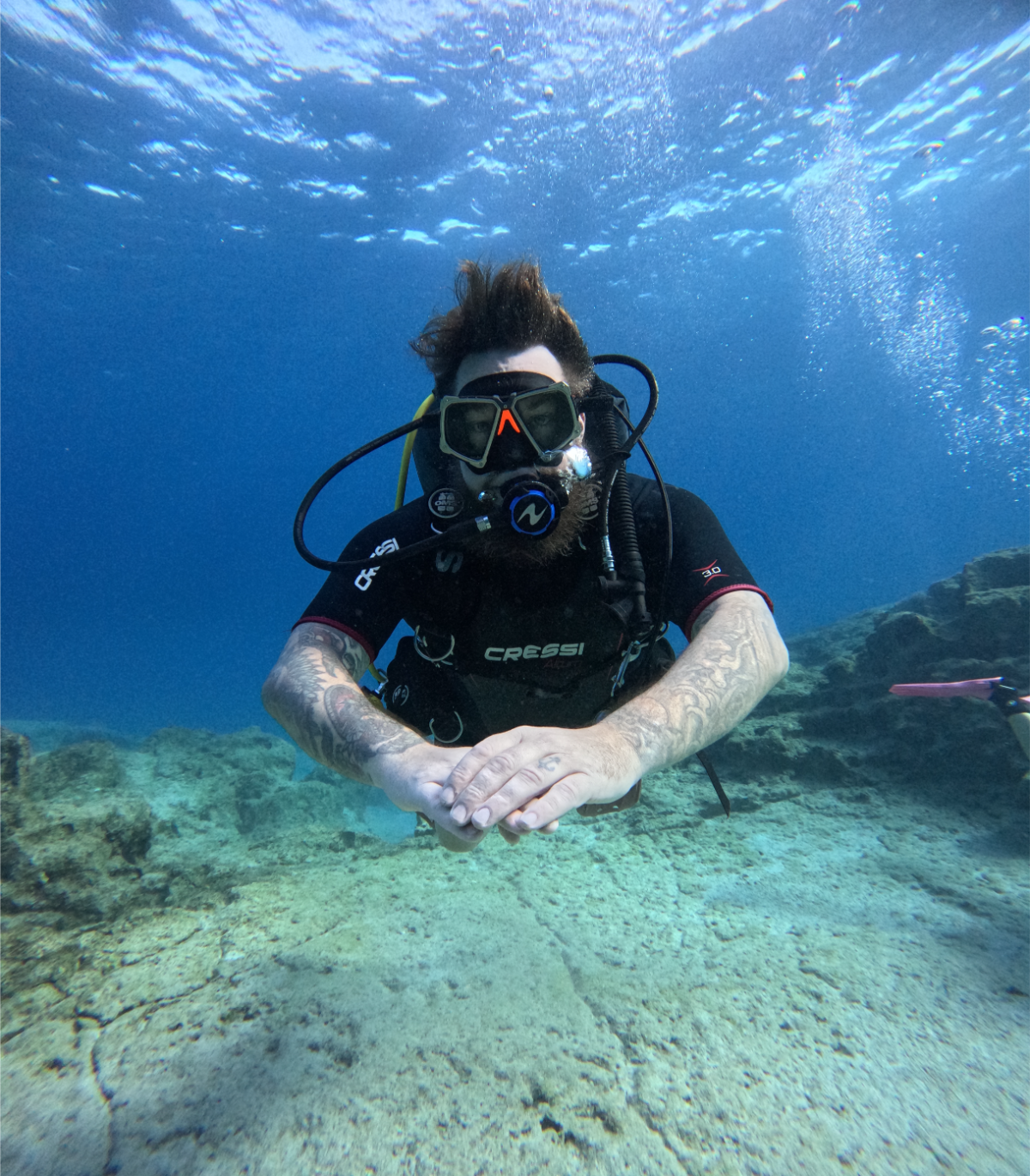 A person scuba diving underwater, wearing a black diving suit and goggles, with arms extended forward, surrounded by blue water and ocean floor rocks.