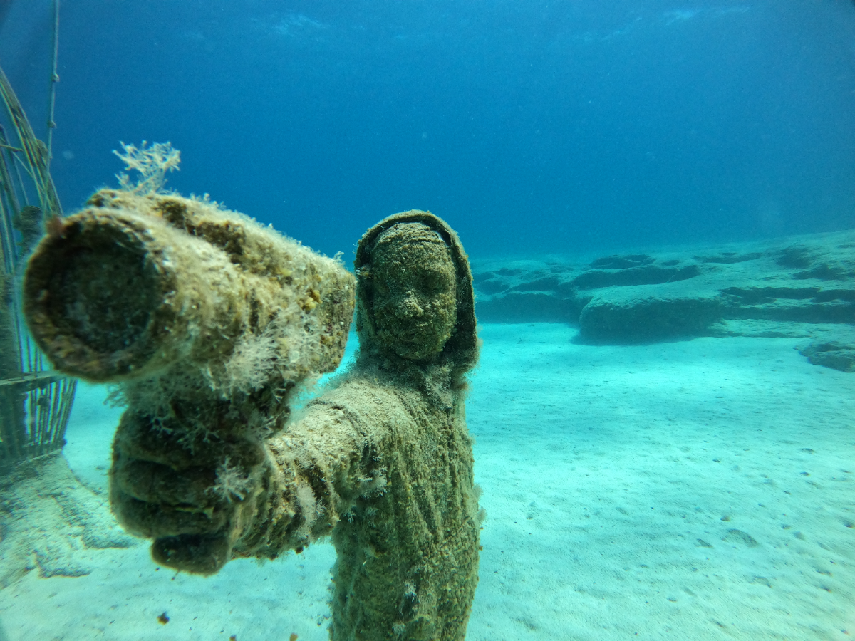 U.S. subject covered in marine growth underwater, with arm extended toward the camera, in a sandy underwater environment.