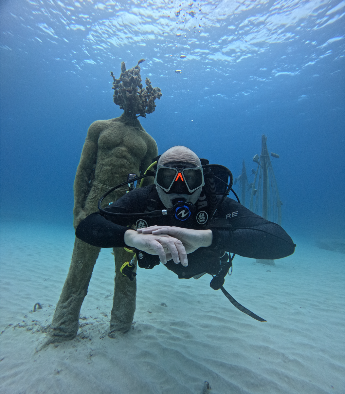 A scuba diver underwater wearing a black wetsuit, mask, and breathing gear, with a humanoid figure statue with a tree-shaped head and moss-covered body in the background at MUSAN.