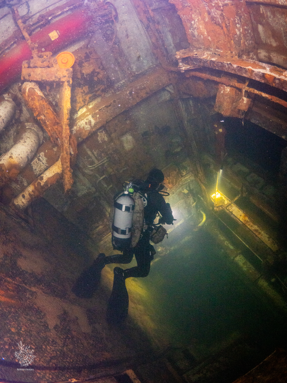A scuba diver swimming inside a submerged, rusted, industrial structure or shipwreck with walls and pipes covered in rust.