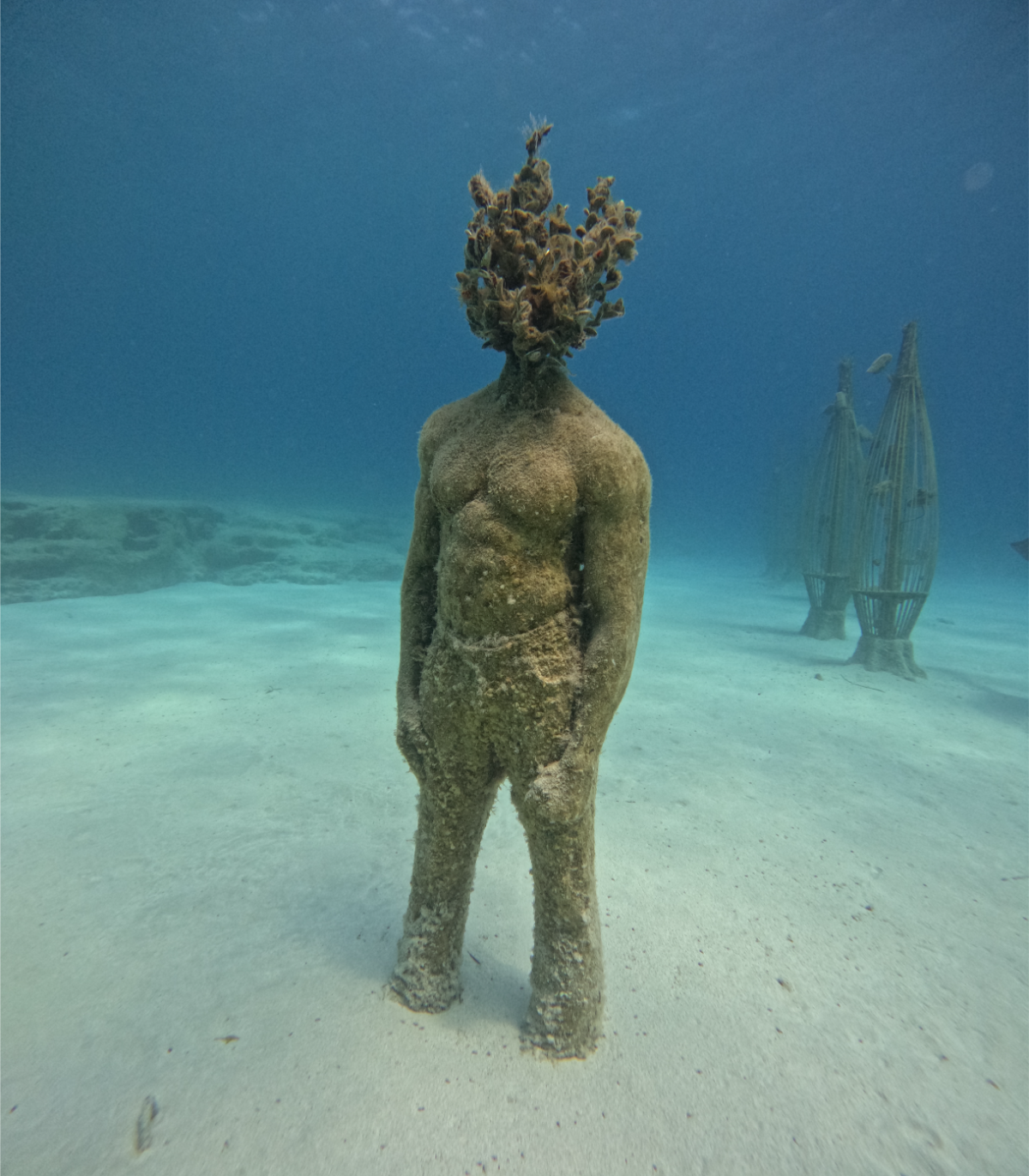An underwater sculpture of a human figure with a body covered in sand or algae, and a head made of coral and marine growth, standing on the sandy ocean floor.