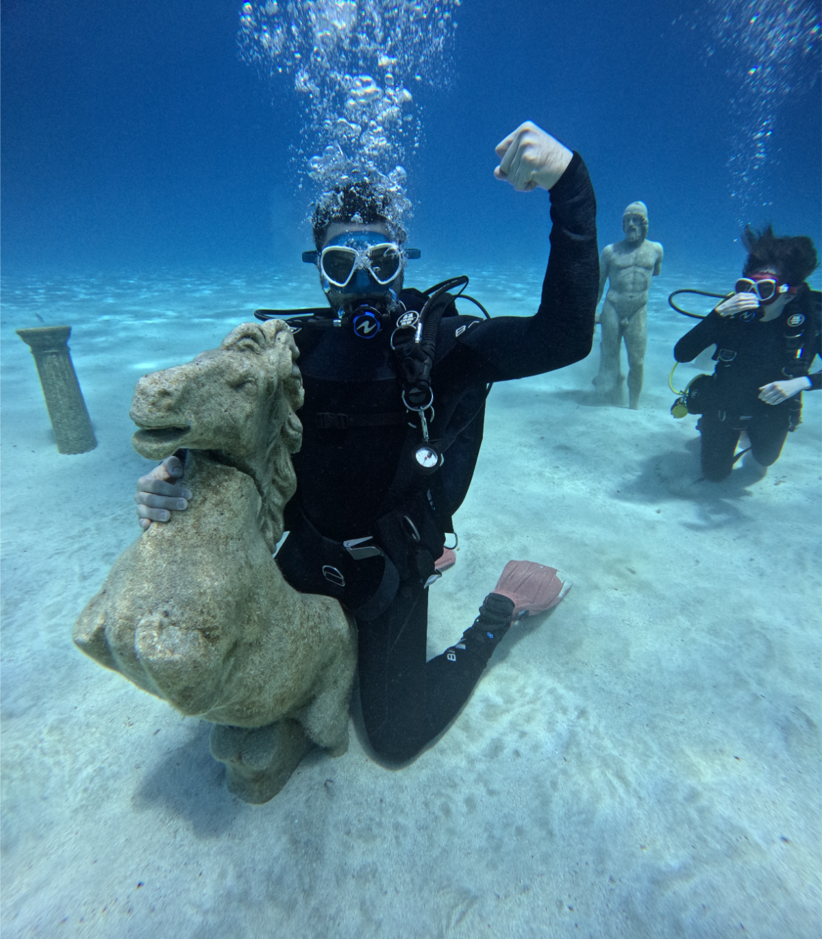 A person in scuba gear underwater kneeling on the ocean floor, flexing their arm with a sculpture of a lion's head. Two other scuba divers are in the background near classical statues, with one person kneeling and the other floating.