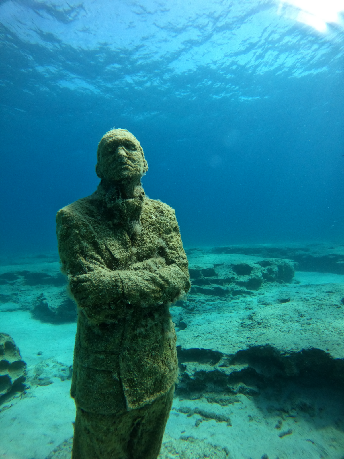 Underwater scene of a statue of a man with his arms crossed, covered in algae and coral, with a thoughtful expression, and rocks on the ocean floor in the background.