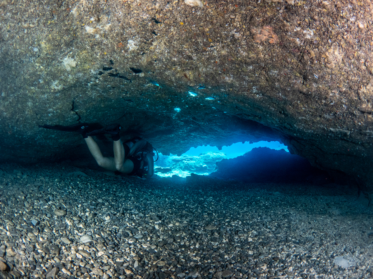 A person in scuba gear crawling through a narrow underwater cave with a rocky floor, illuminated by blue light from outside.