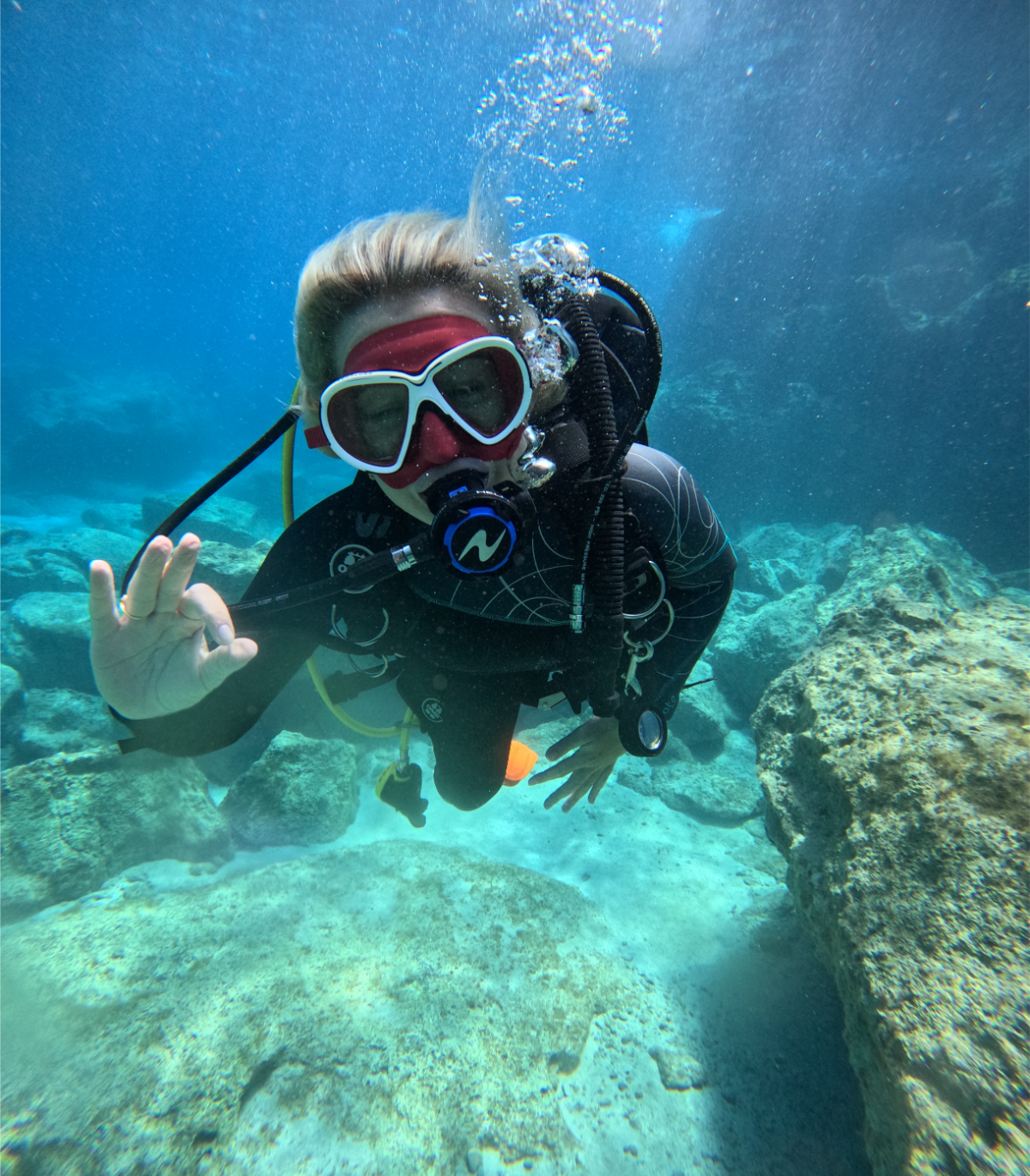 A woman scuba diving underwater, wearing a diving mask, snorkel, and wetsuit, making an OK gesture with her hand while surrounded by coral and rocks.