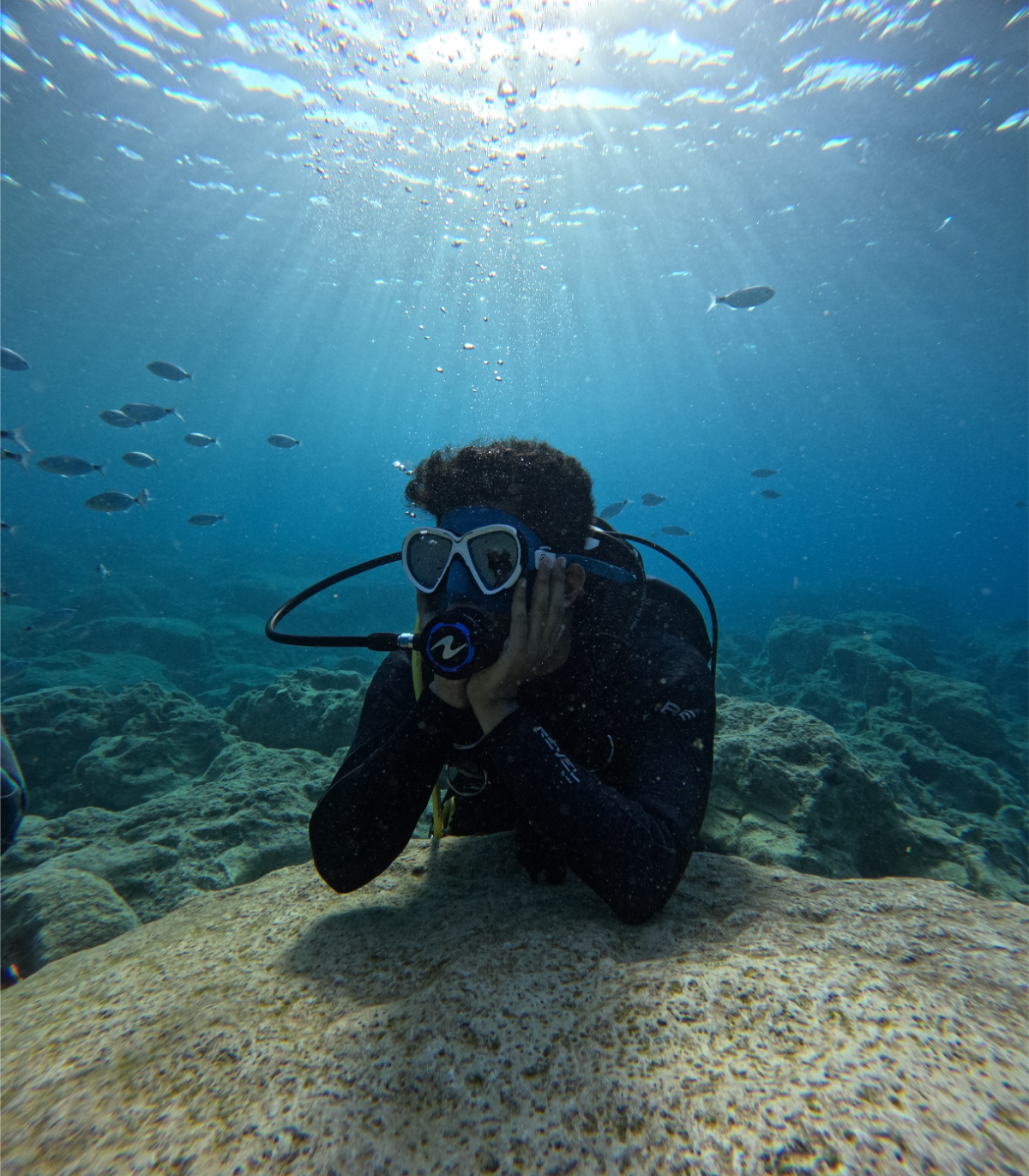 Person scuba diving underwater with hand on cheek, wearing snorkeling gear, surrounded by fish and coral reef, with sunlight filtering through the water