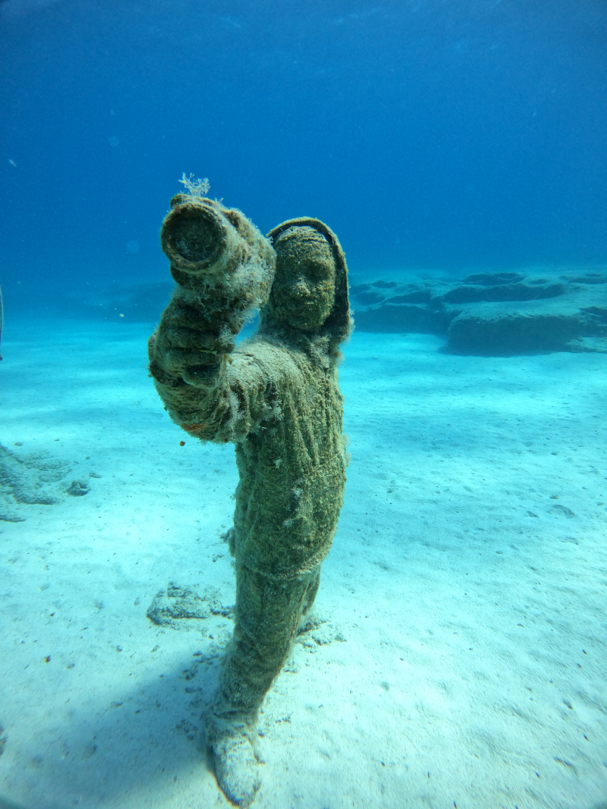 Underwater sculpture of a girl with a scuba snorkel, covered in algae and marine growth, standing on the ocean floor with a sandy seabed and rocks in the background.