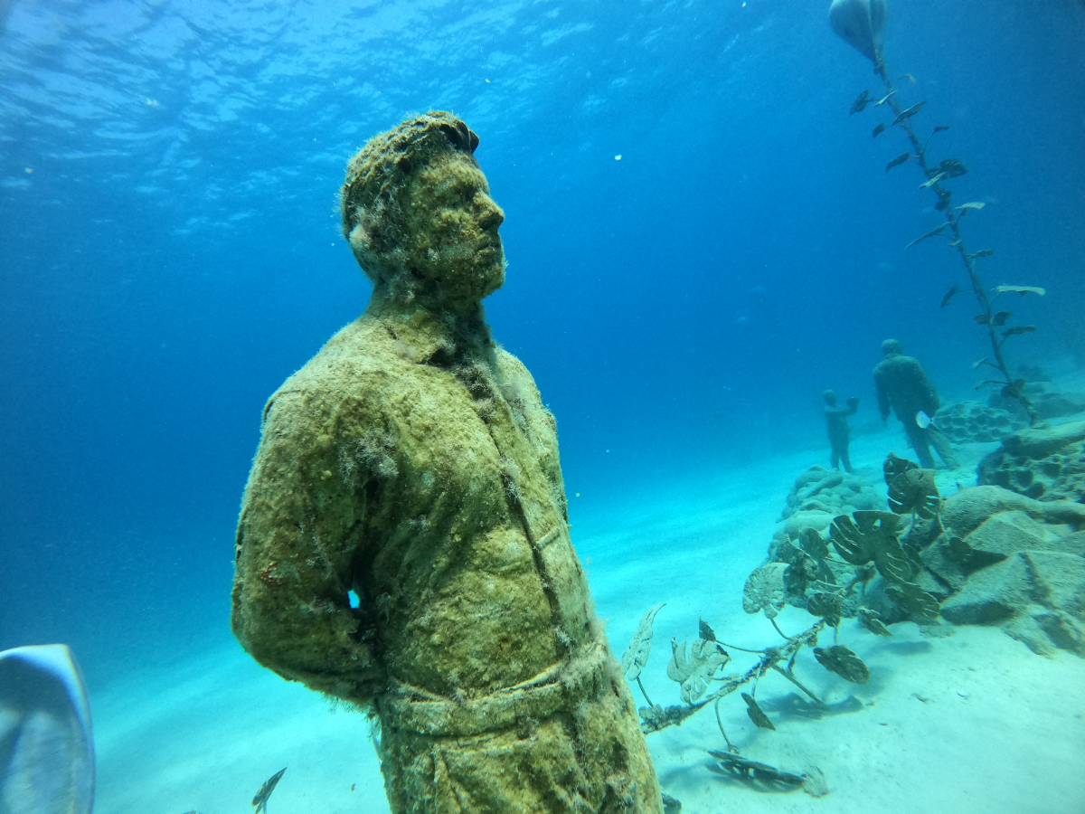 An underwater statue of a man in a coat and scarf, covered in algae and marine growth, with a group of divers in the background exploring a shipwreck or reef.