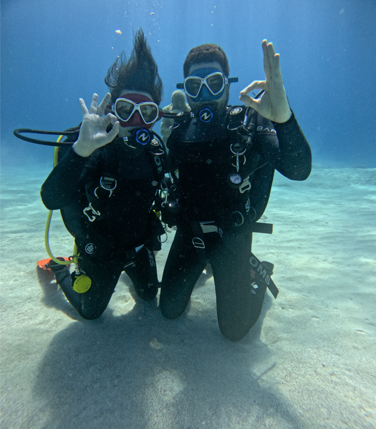 Two scuba divers kneeling on the sandy ocean floor, wearing wetsuits, masks, and scuba gear, making hand signals underwater.