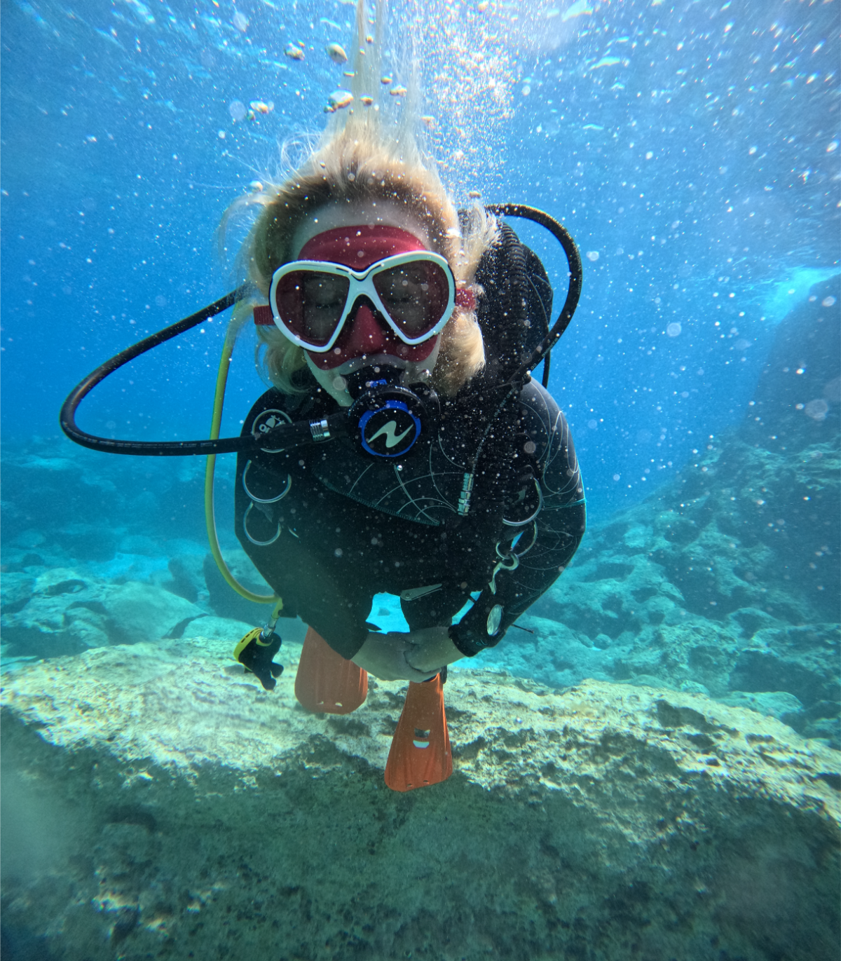 Person scuba diving underwater, wearing mask, wetsuit, and fins, surrounded by blue water and underwater rocks.