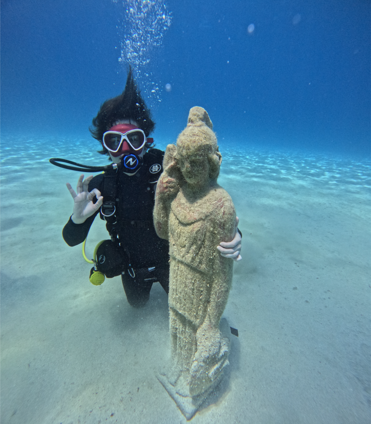 A scuba diver underwater holding a stone statue at Green Bay, making an OK sign with their hand.